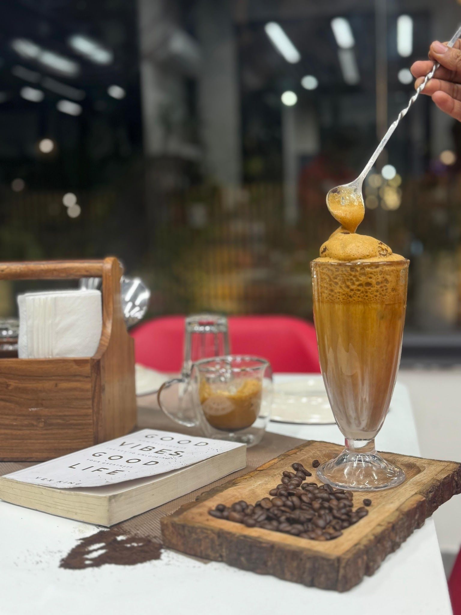 A tall glass of whipped iced coffee being stirred beside coffee beans and a book